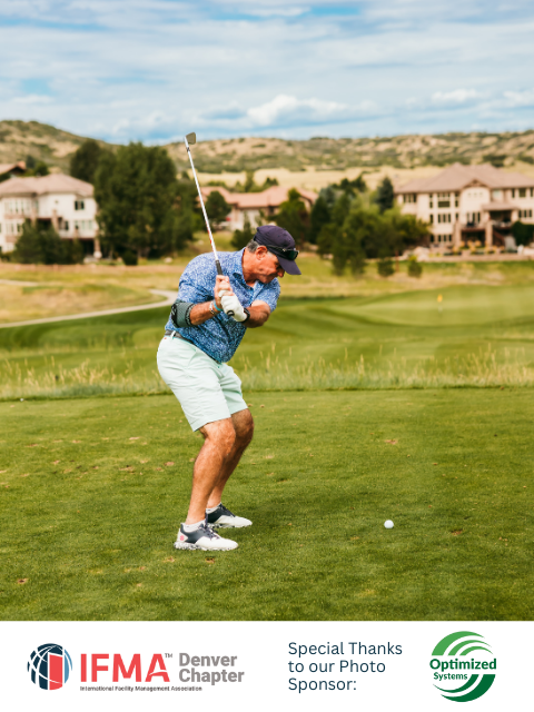 Man swinging a golf club on a green, with golf course and buildings in the background.