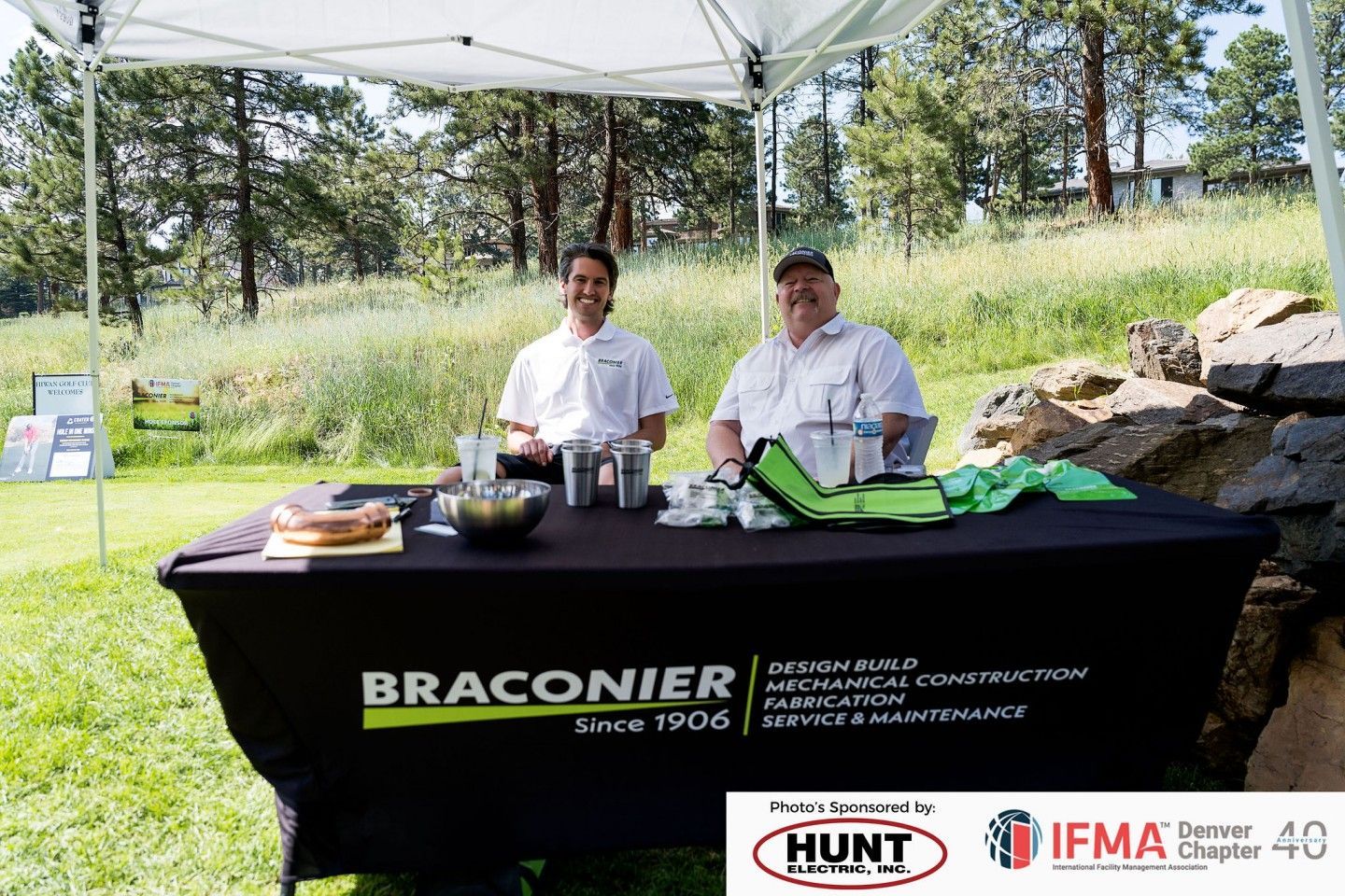 Two men at a Braconier booth outdoors, under a tent. They're smiling, behind a table with items, sponsored by Hunt and IFMA.