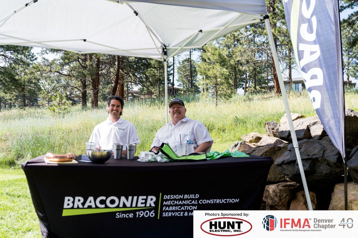 Two men at a Braconier Mechanical Construction booth under a white tent; table with company logo.