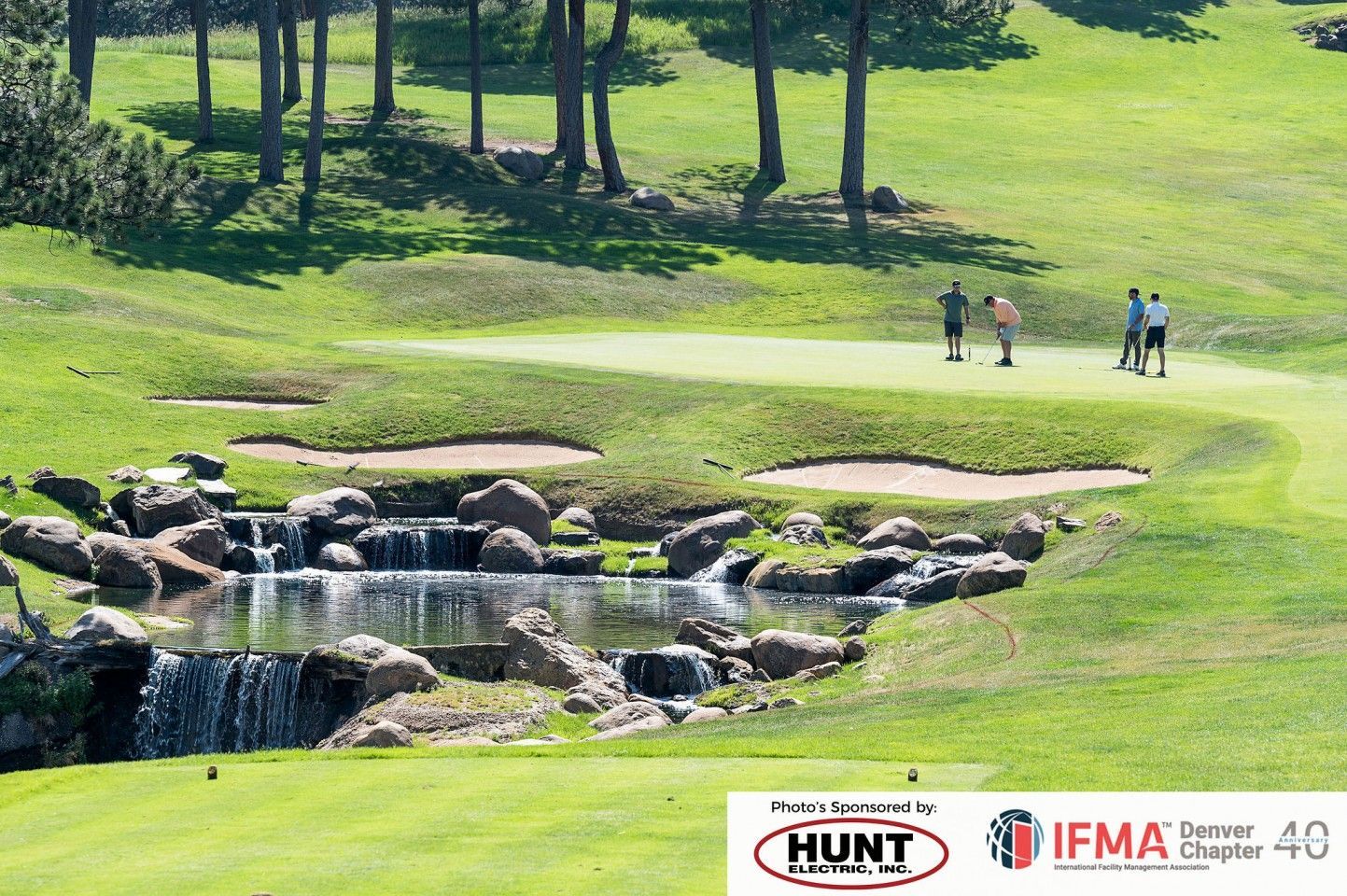 Golfers near a green, with a water feature and sand traps. Green grass, trees, sunny day.