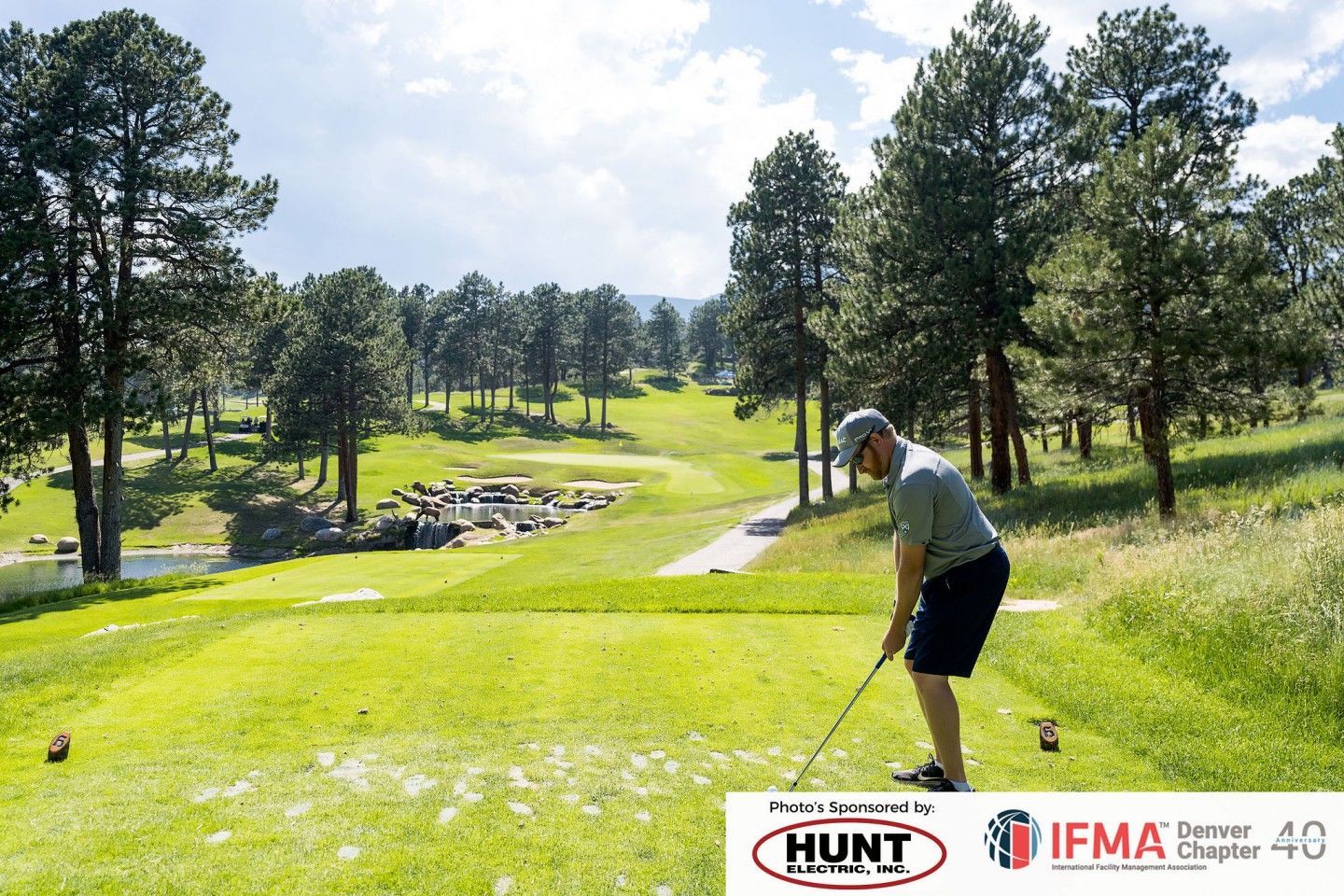 Golfer teeing off on a green golf course, surrounded by trees under a bright sky.