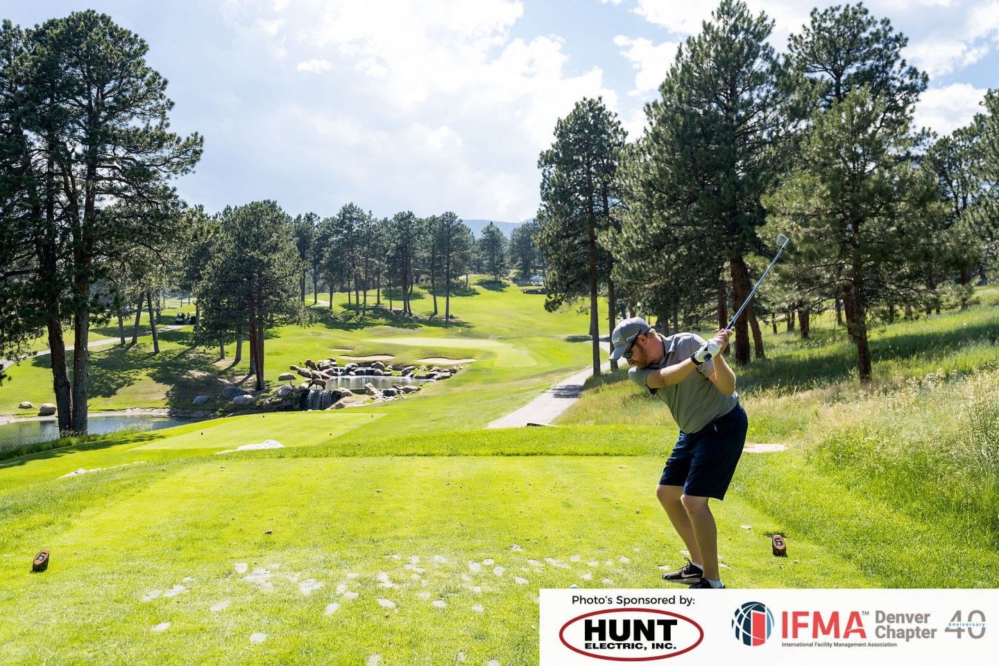 Golfer tees off on a green golf course, trees in the background, under a bright sky.