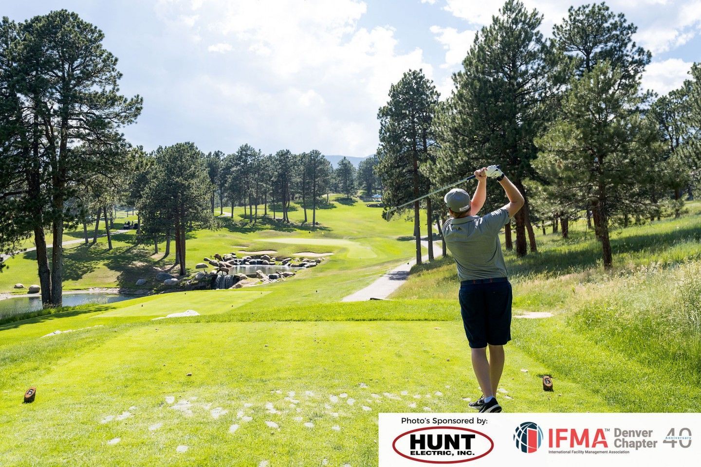 Golfer teeing off on a sunny golf course, surrounded by trees and a pond.