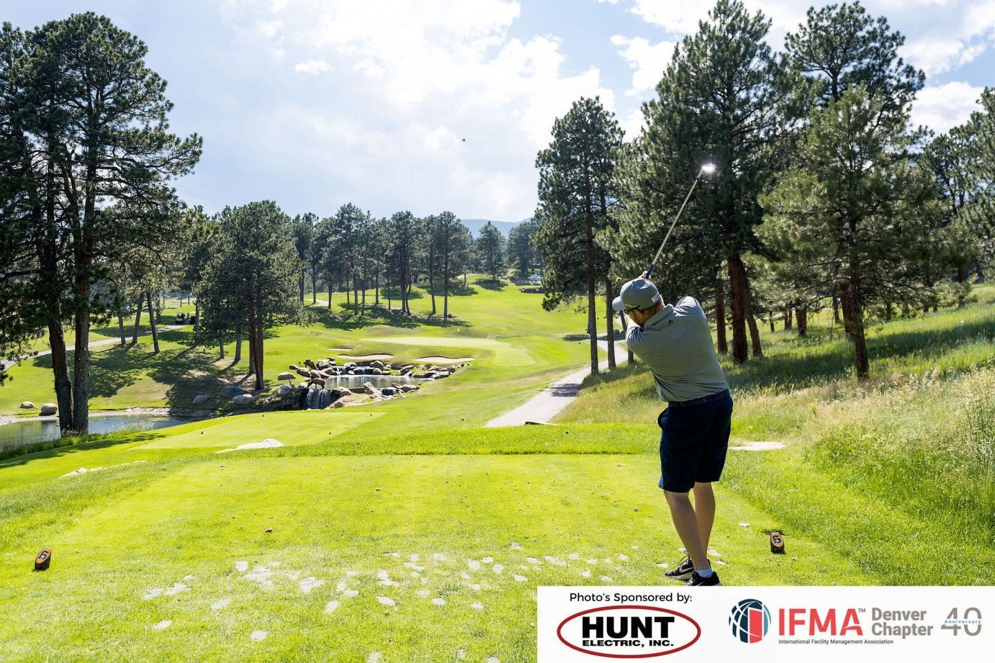 Golfer swings club on a green golf course, tall trees, cloudy sky.