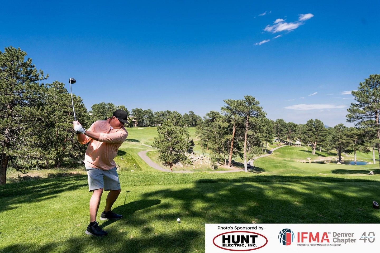 Man swings a golf club on a green course under a blue sky, trees in the background.