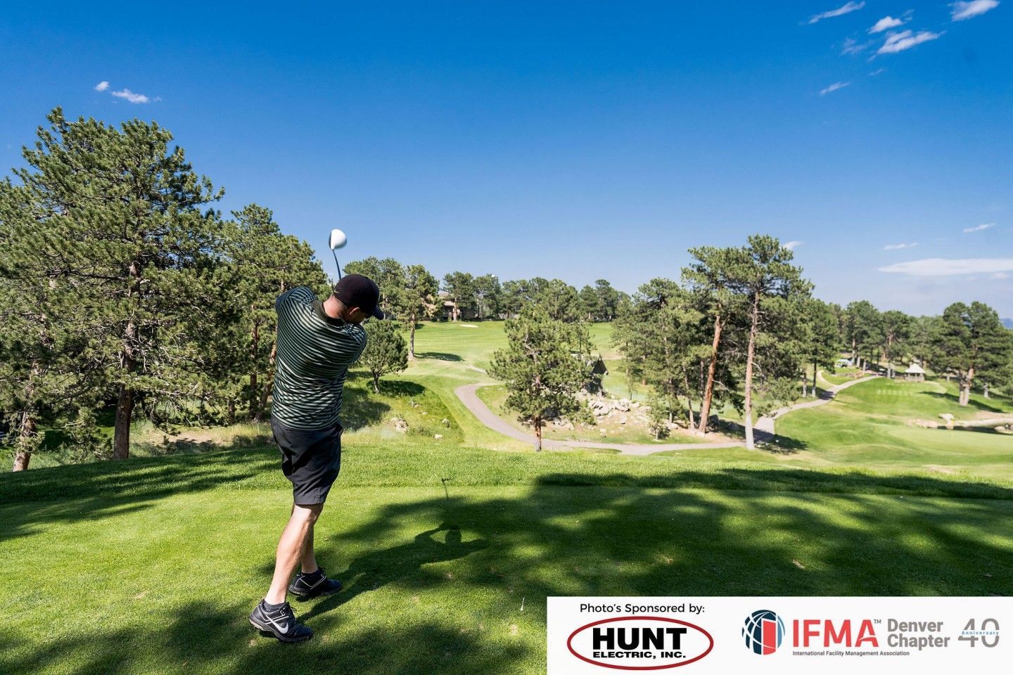 Man swinging golf club on a green course under a bright blue sky.