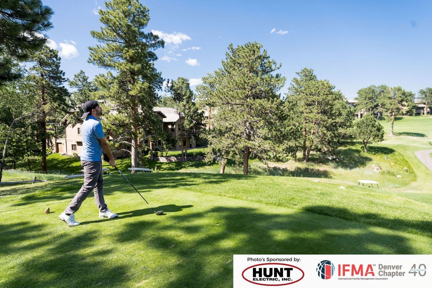 Man walking on a golf course, holding a golf club. Trees and buildings are in the background. Sunny day.