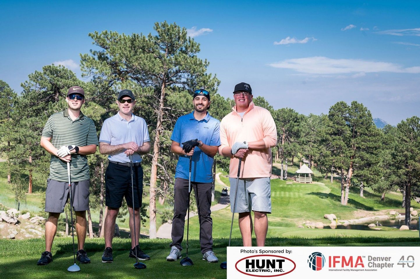 Four men on a golf course holding clubs, posing for a photo. Lush green landscape, sunny day.