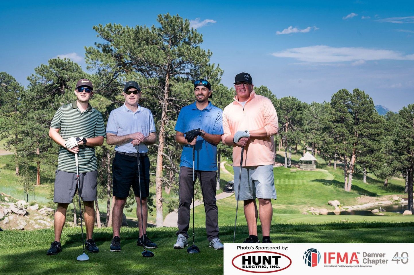 Four men on a golf course holding clubs, posing for a photo. Blue sky and trees in the background.