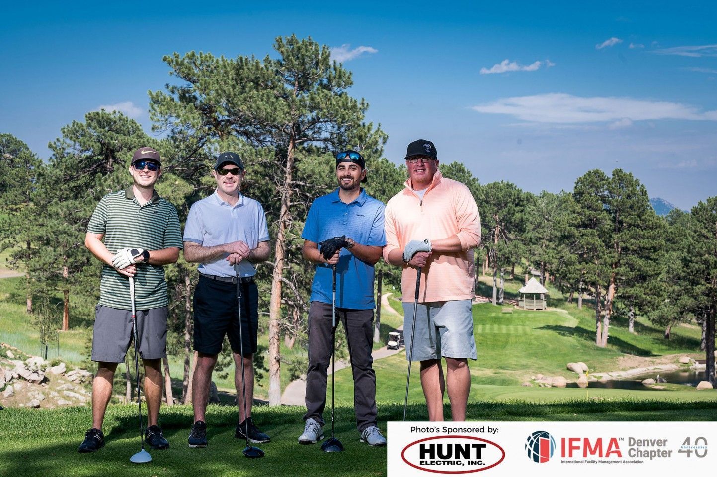 Four men on a golf course holding clubs, posing for a photo. Blue sky, green grass.