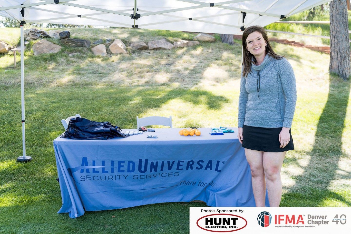 Woman stands near a table with Allied Universal Security Services logo under a tent on grassy lawn.