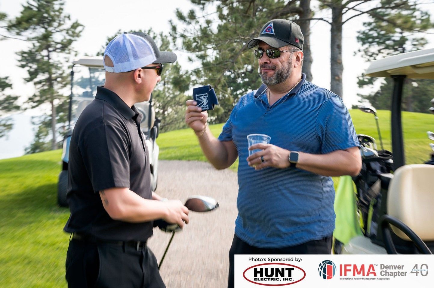Two men talking on a golf course, one holding a hat, another holding a drink. Golf carts are nearby.