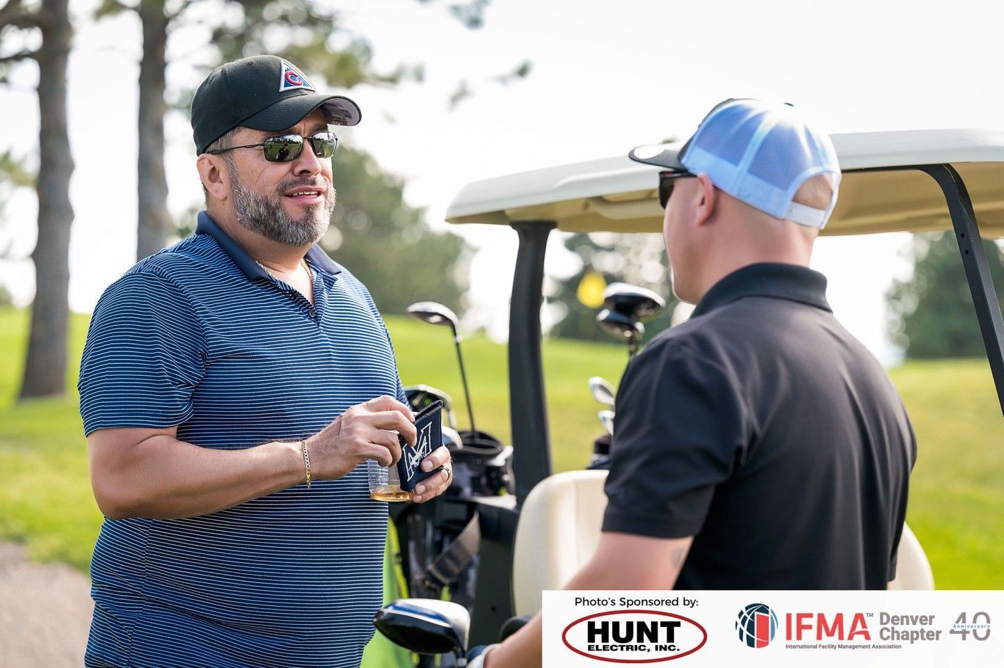 Two men in sunglasses talking near a golf cart on a course. One holds a beverage.
