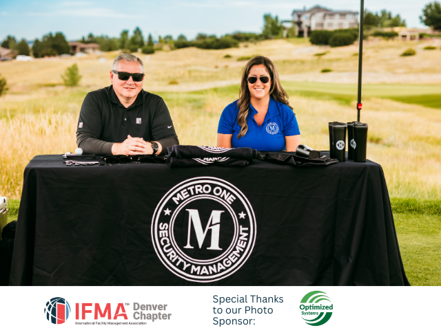 Two people at a Metro One Security Management table at an outdoor event, with logo, golf course background.