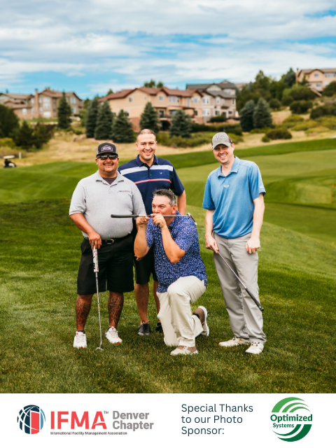 Four men pose on a golf course. One kneels and makes a funny face. Buildings and trees are in the background.