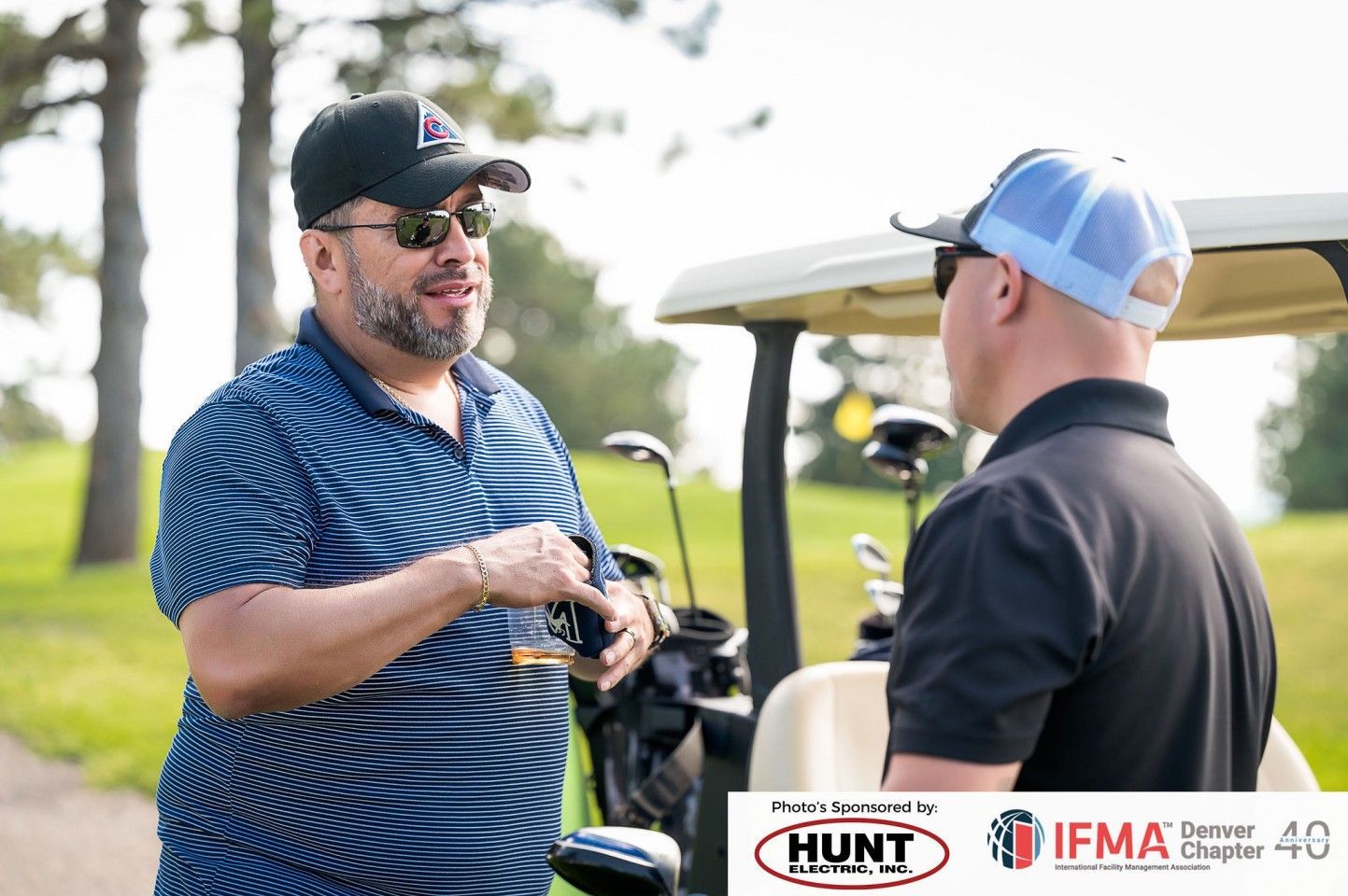 Two men talking by a golf cart on a green course; one gestures while holding a glass.