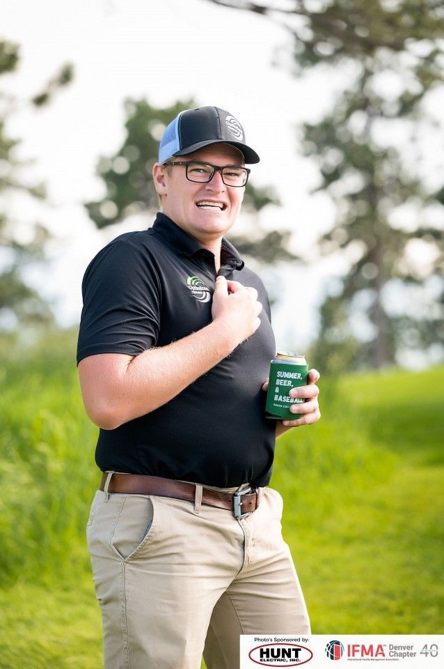 Man in black shirt and hat, holding a drink, smiling outdoors.