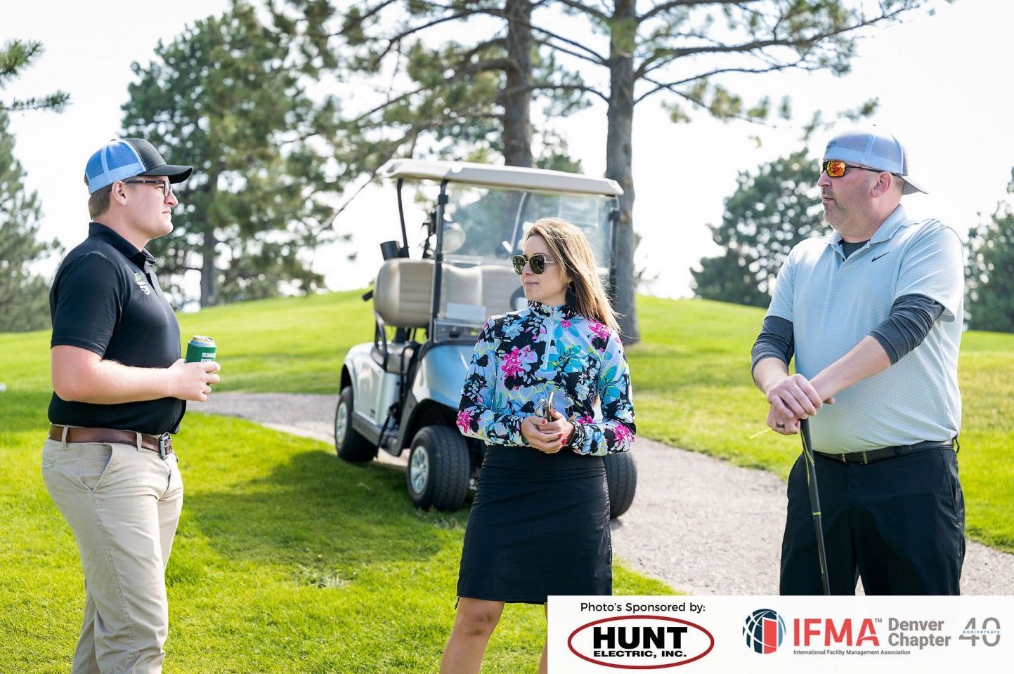 Three people on a golf course with a golf cart. One holding a drink, two others holding golf clubs.