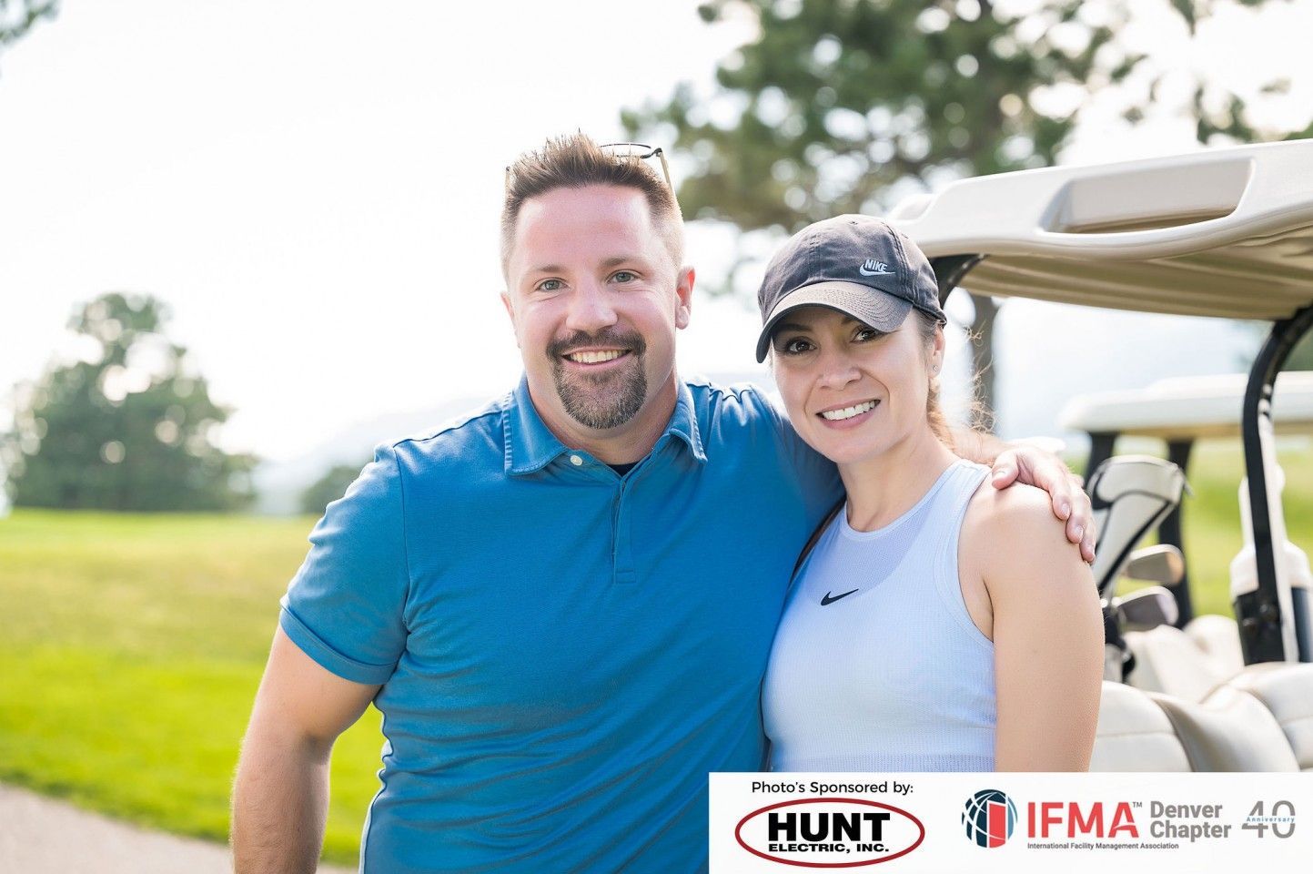 Man in blue shirt and woman in cap smile, posing by golf cart. Outdoor setting.