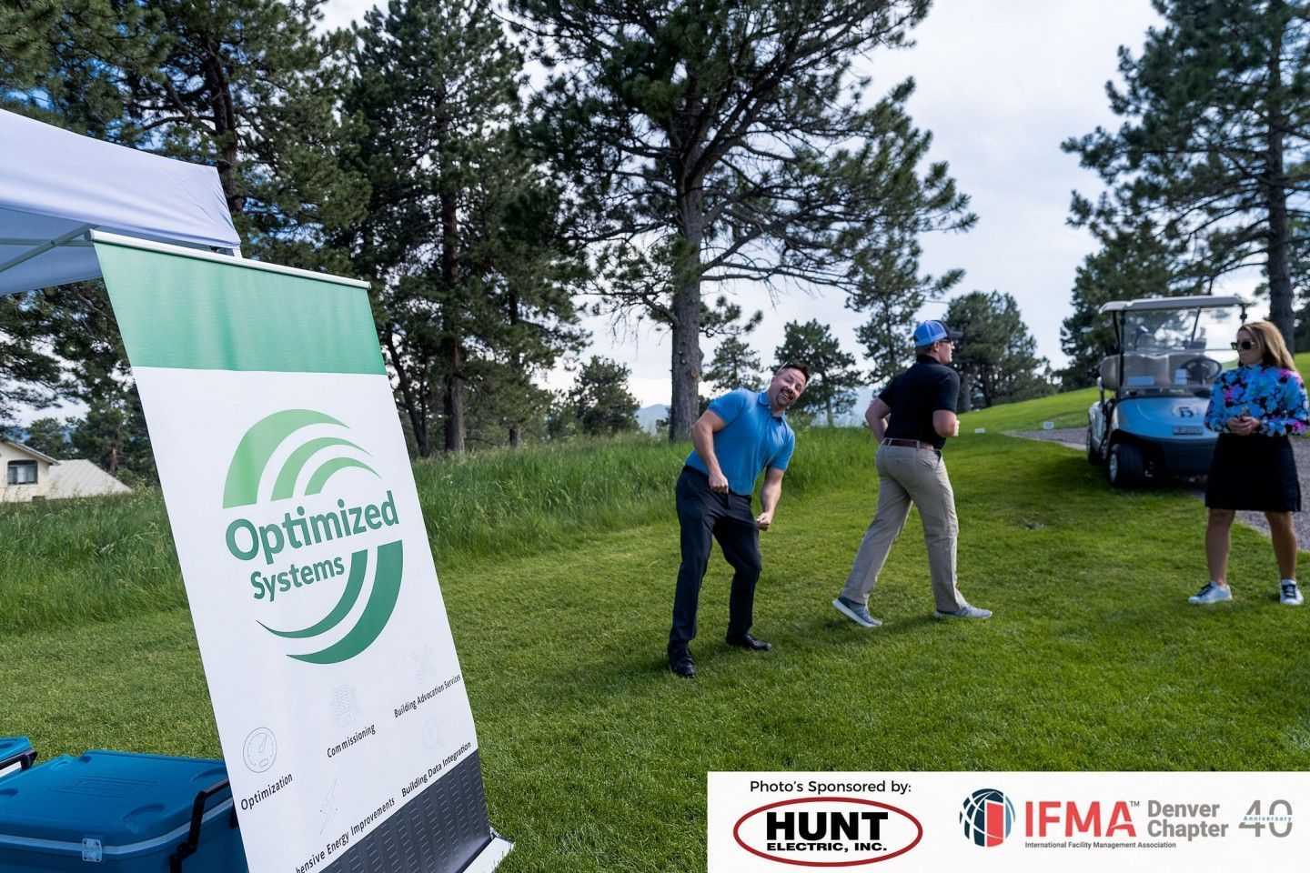 Golf tournament with Optimized Systems banner. Two men golfing; cart and woman nearby. Outdoors, green field.