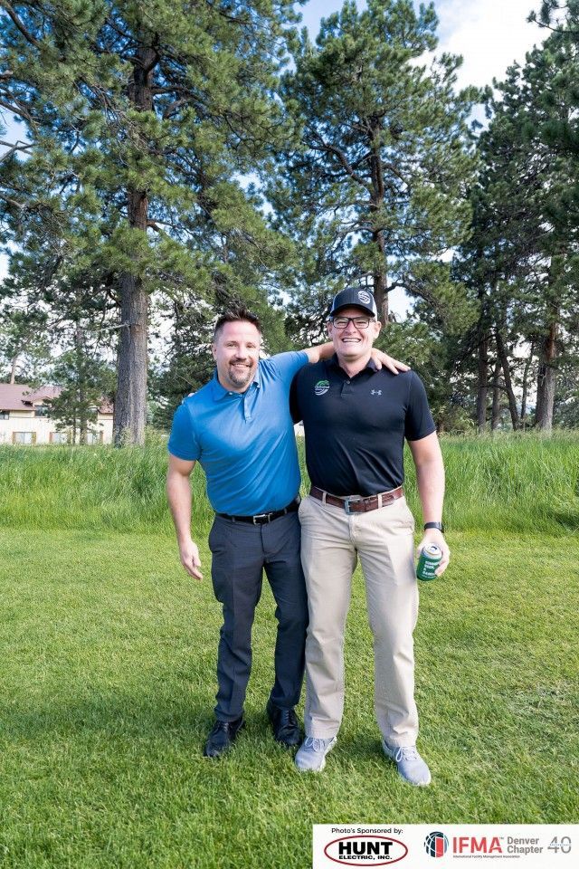 Two men smiling, posing on a golf course. One has arm around the other's shoulder. Green grass and trees in background.