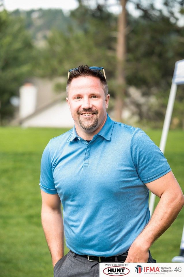 Man in blue polo shirt smiles, posing outdoors. Sunglasses atop head, hands in pockets.