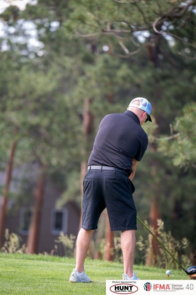 Golfer in black shirt and shorts swings at a golf ball on a green. Trees in the background.