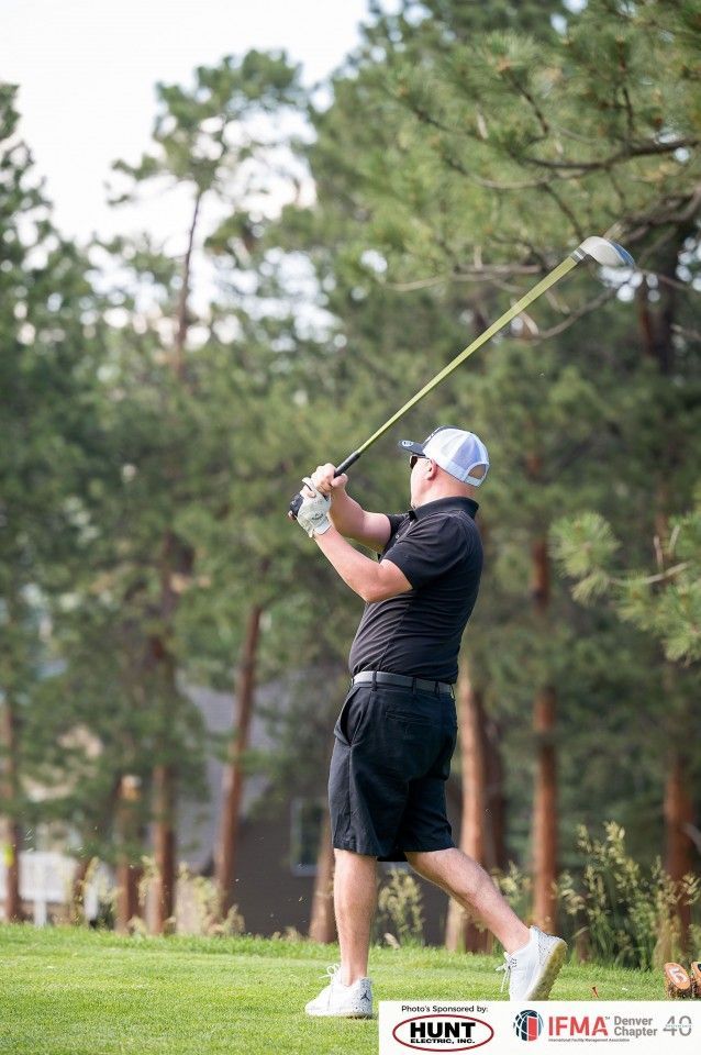 Man swinging a golf club on a green course, trees in the background.