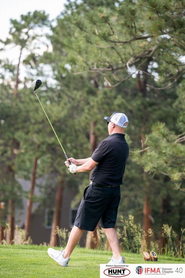 Golfer swings club at a golf course. Wearing black shirt, shorts, and a hat. Green grass and trees are in the background.