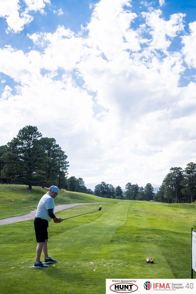 Man teeing off on a golf course. Green grass, blue sky with clouds, trees in the background.