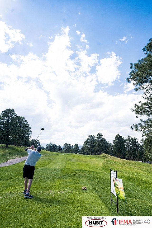 Golfer swings at tee on a green fairway under a partly cloudy sky. Trees and a sign on the right.