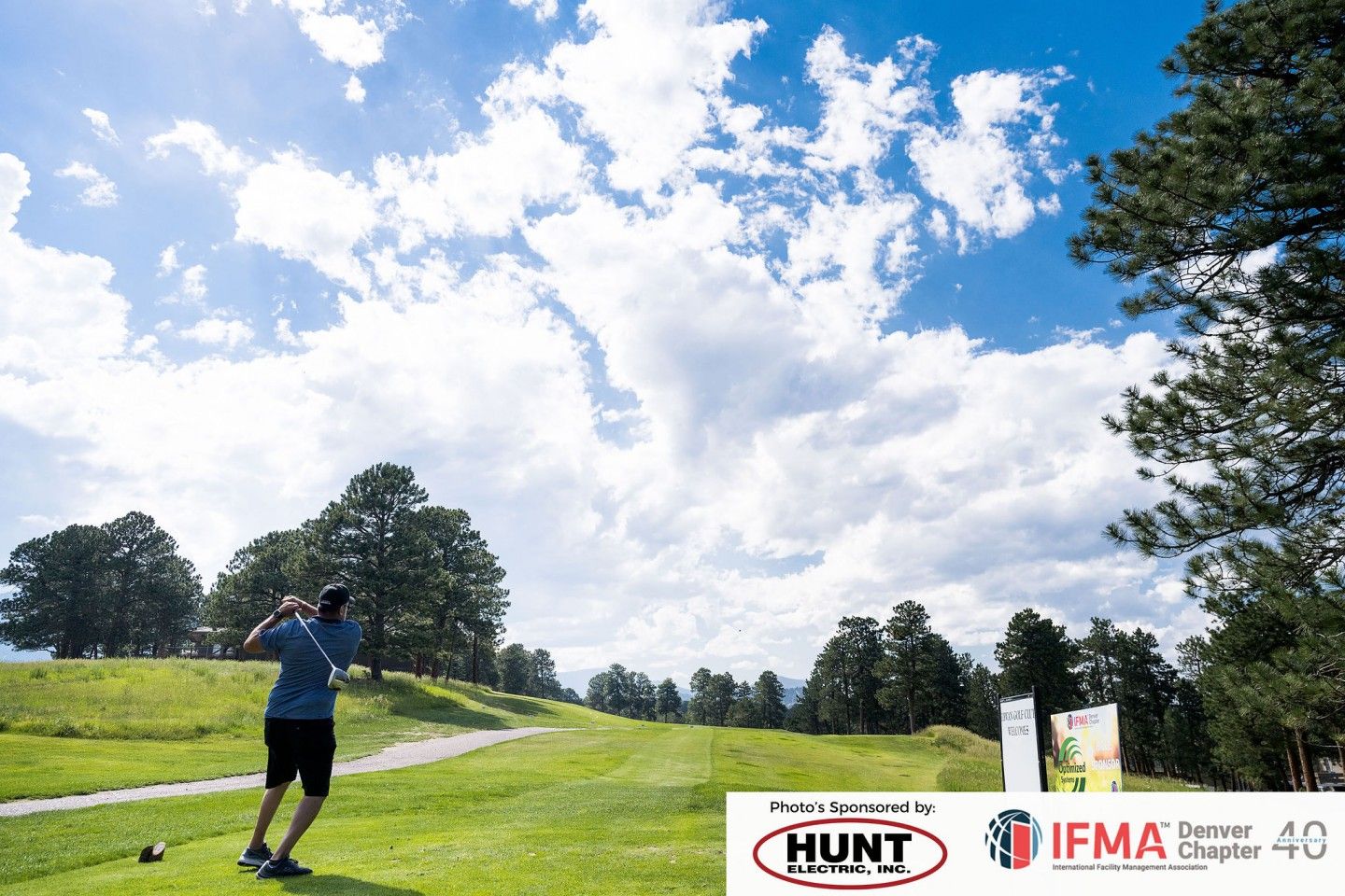 Golfer teeing off on a green golf course under a partly cloudy blue sky. Hunt sign visible.