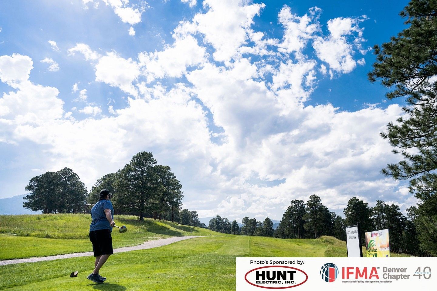 Man tees off on a golf course under a cloudy blue sky. Trees and green grass surround him.