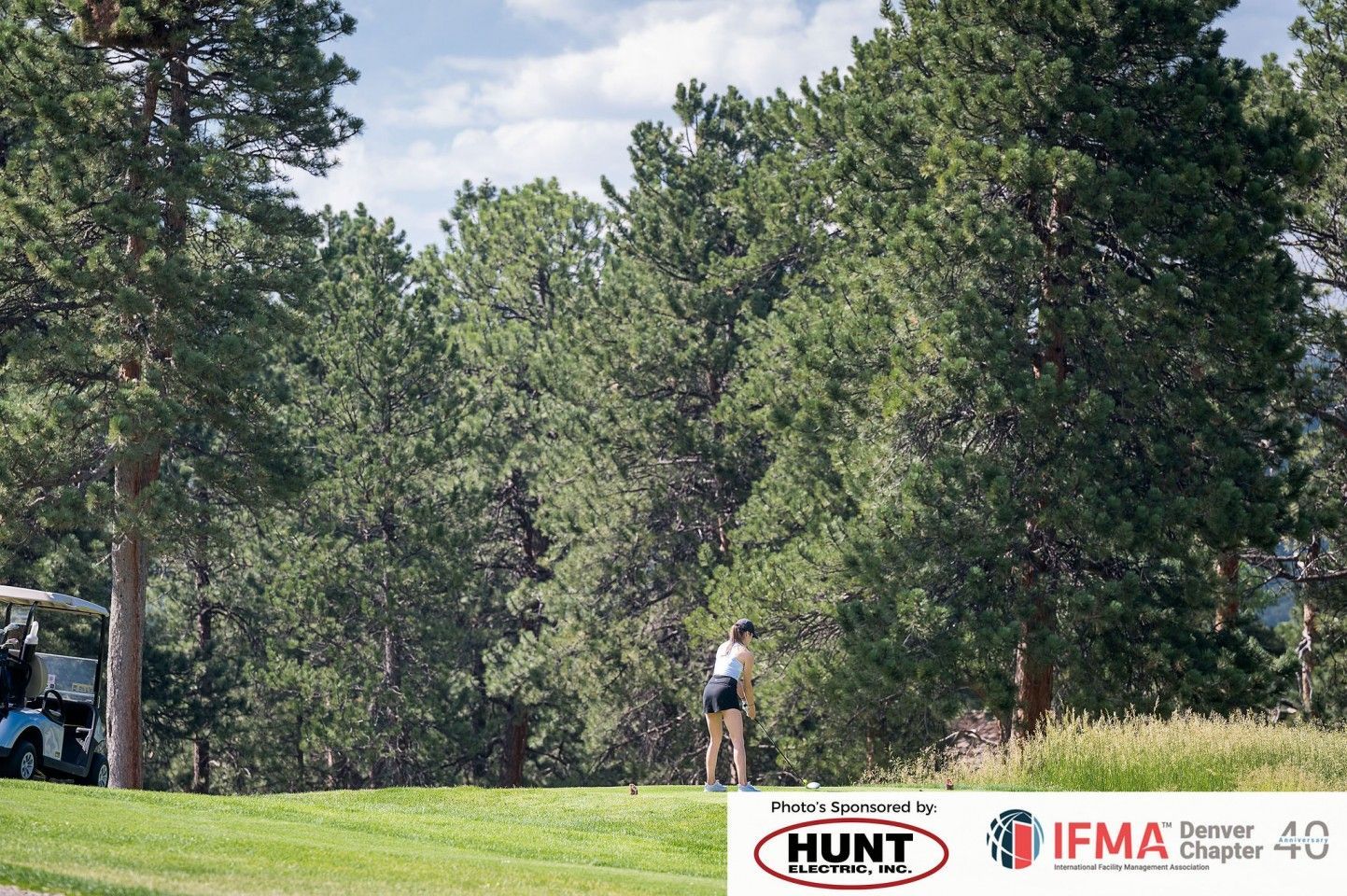 Golfer swings club on a green course, trees in the background, blue golf cart to the left.