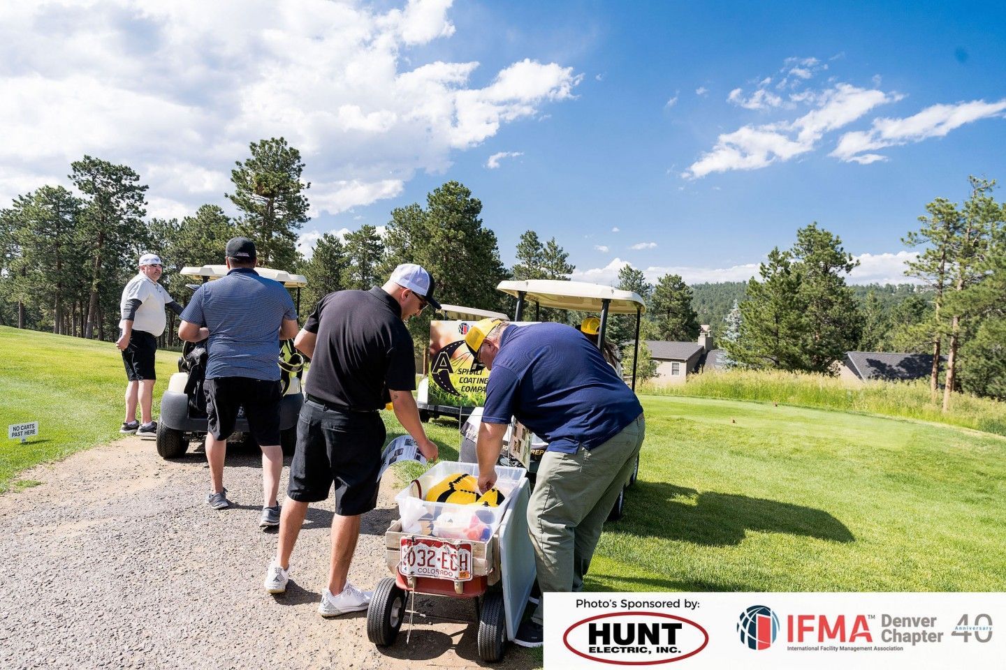 Golfers on a sunny course loading golf carts; trees and blue sky background.