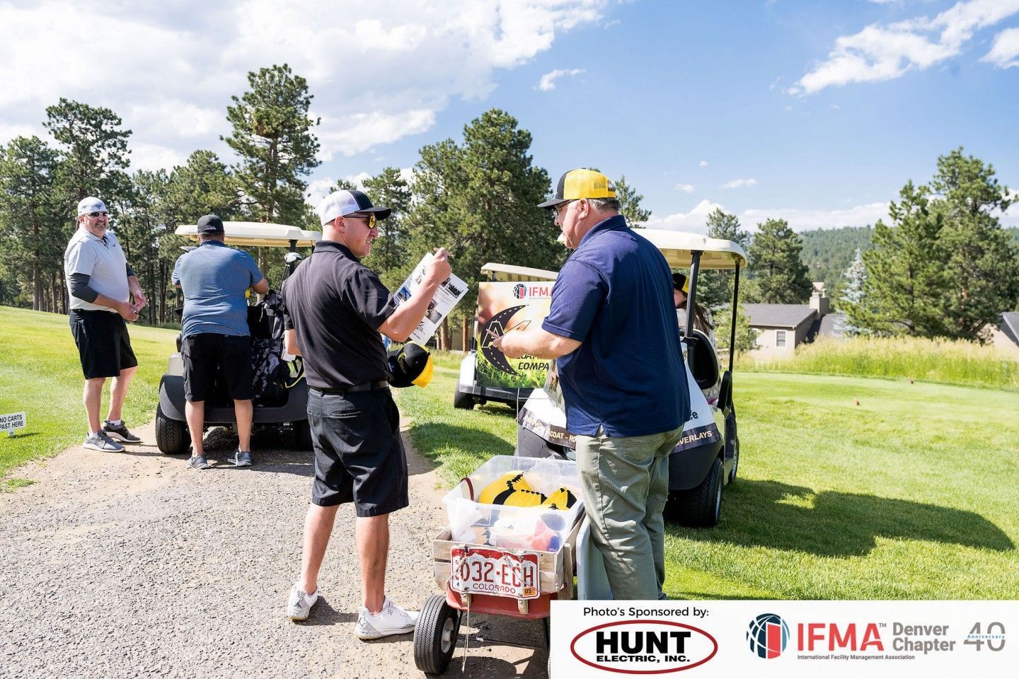 Men on a golf course. One hands a paper to another near a golf cart and wagon of items.