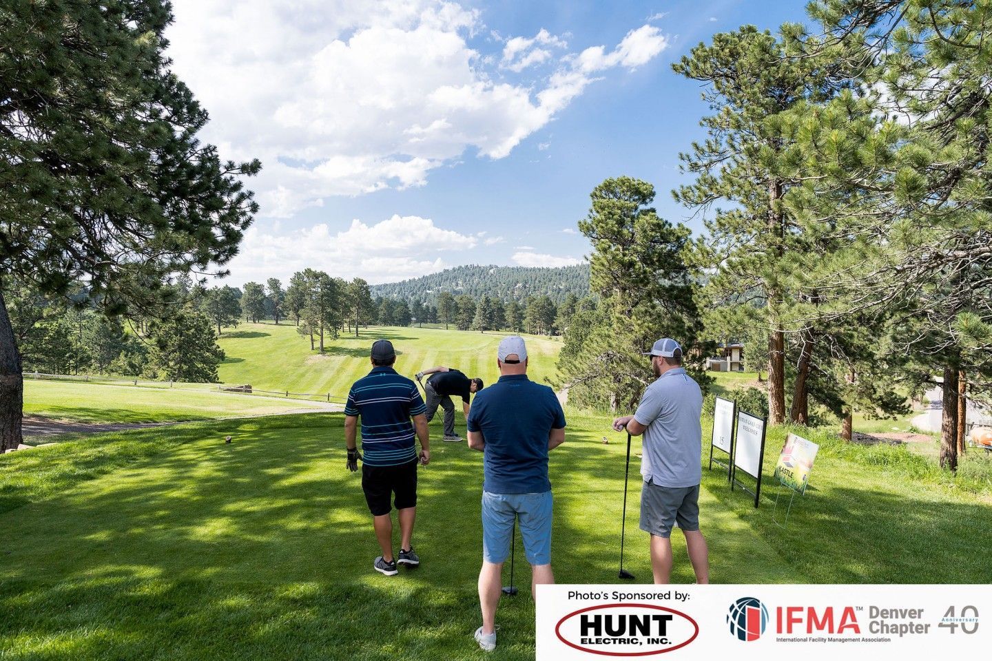 Three golfers on a green golf course, trees and mountains in the background under a blue sky.