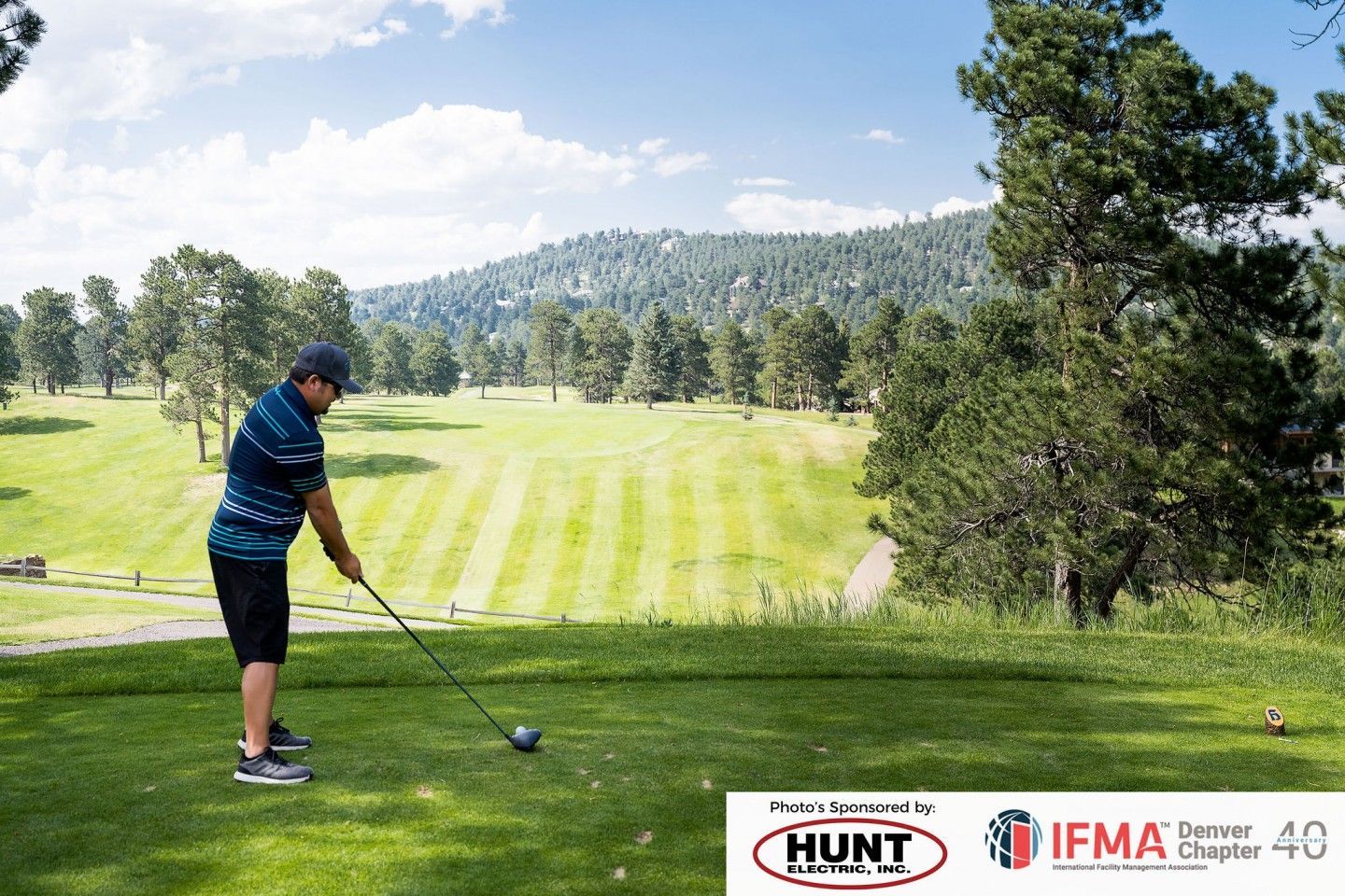 Golfer teeing off on a green golf course, trees in the background, blue sky.