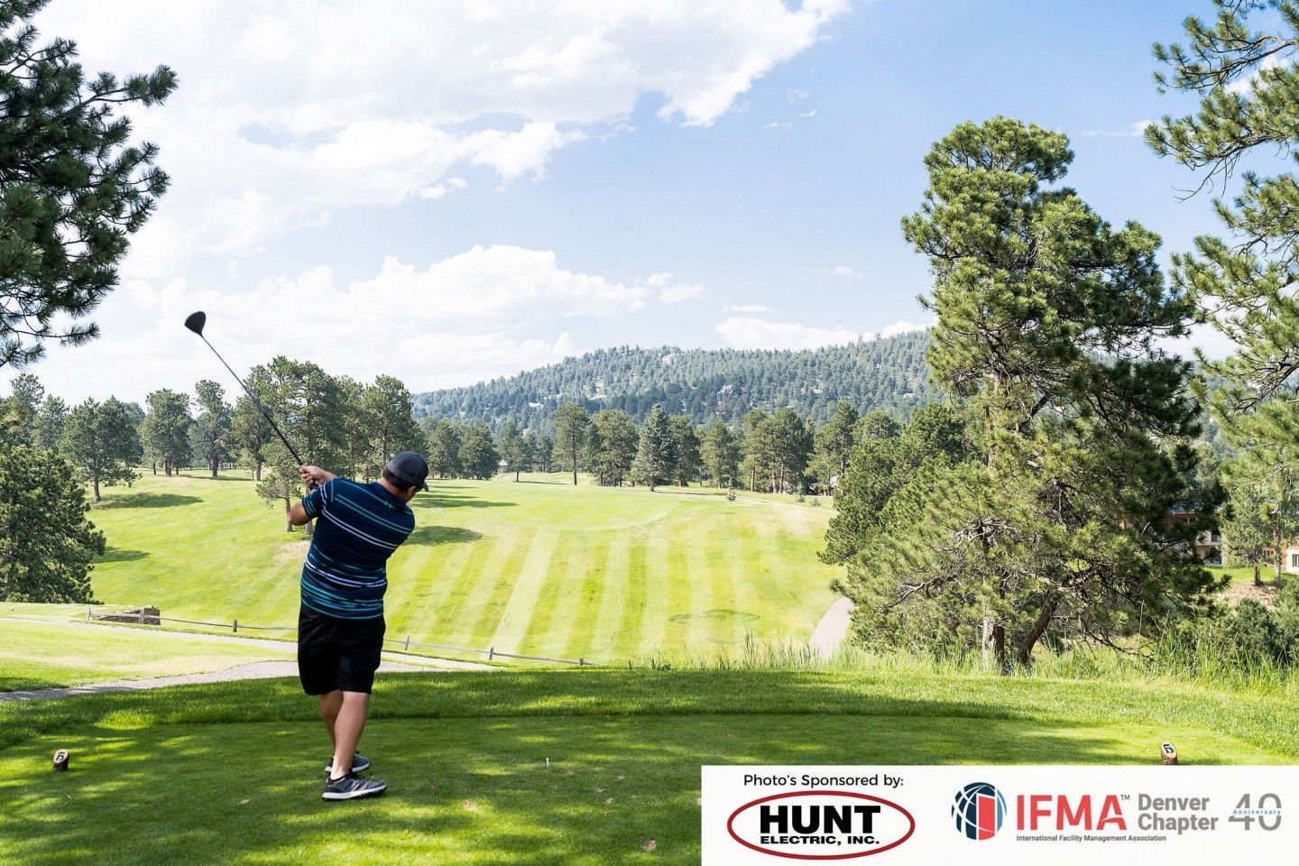 Golfer swings club on a green course with trees, under a blue sky.