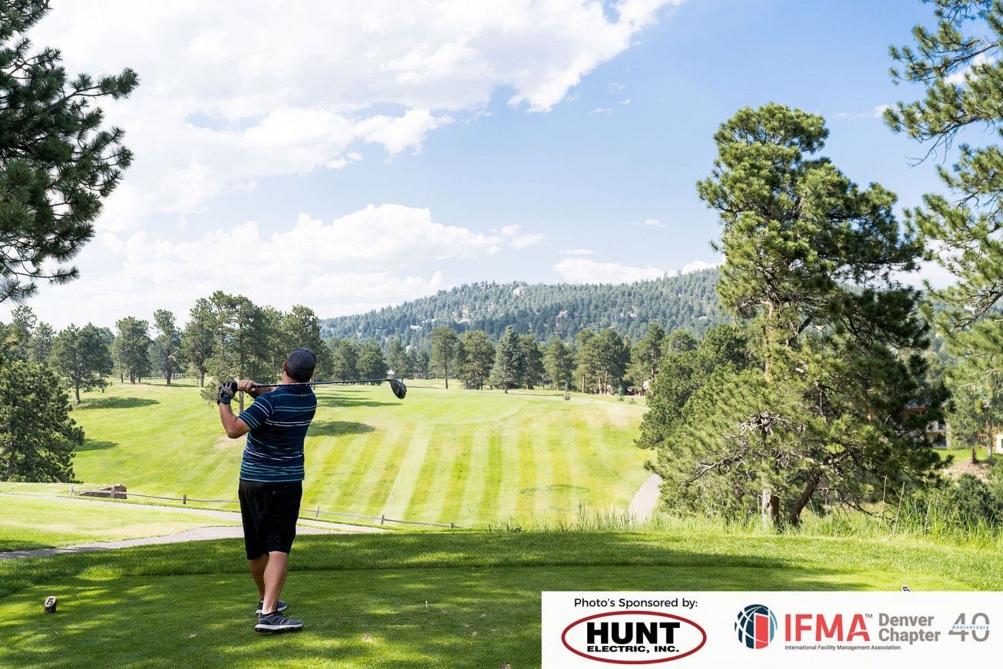 Golfer teeing off on a green golf course, surrounded by trees and mountains under a blue sky.