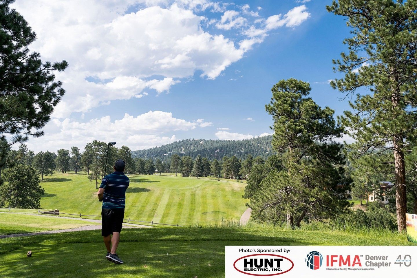 Golfer teeing off on a green golf course under a blue sky, surrounded by trees and a distant hillside.