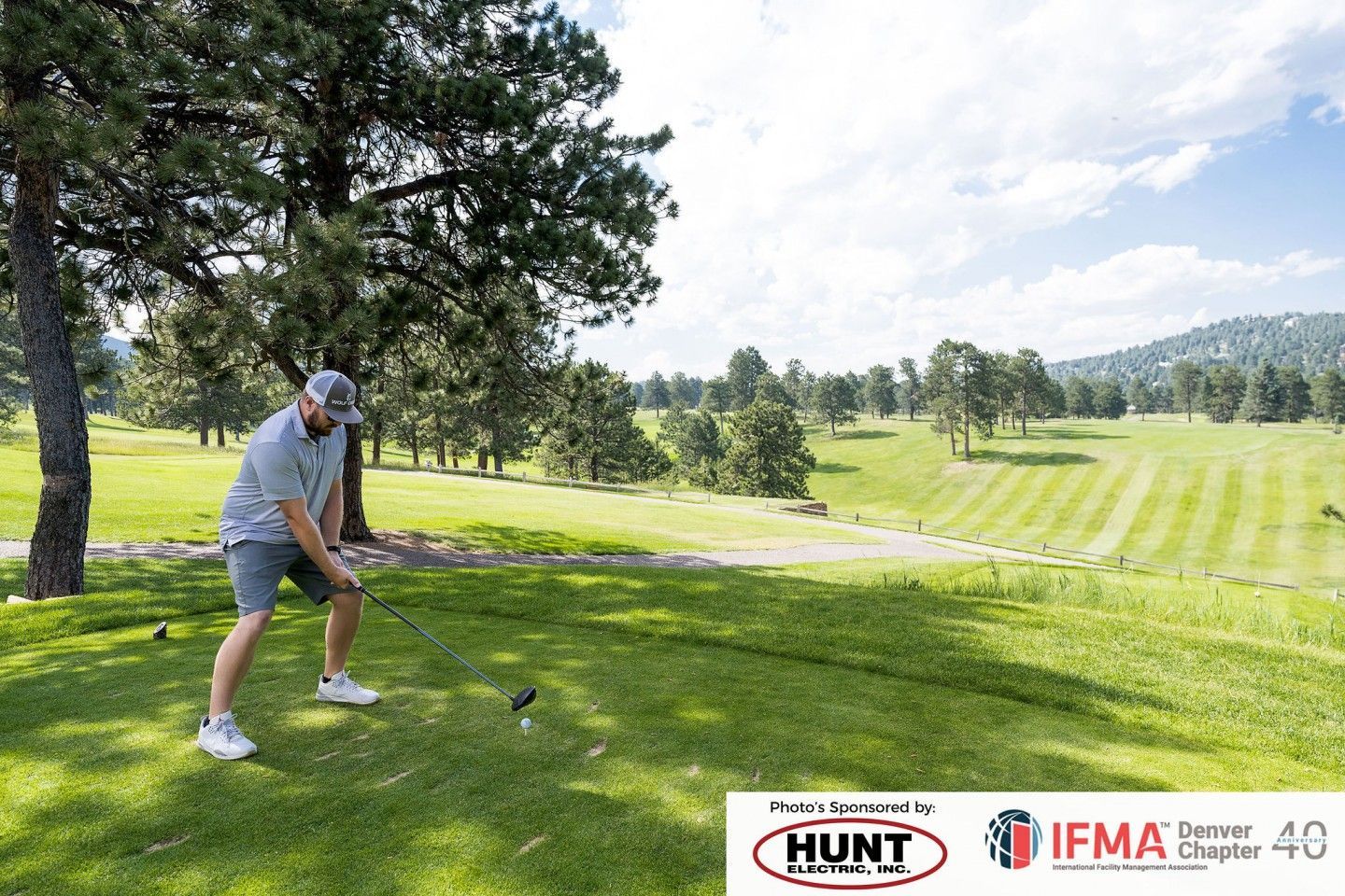 Man swings golf club on a tee, green grass, trees, and distant hills.