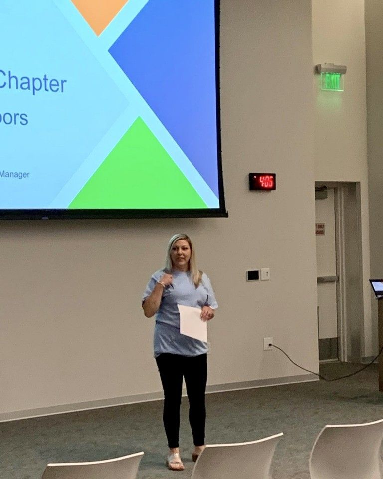 Woman presenting in a lecture hall, pointing at her chest. Presentation screen in the background.