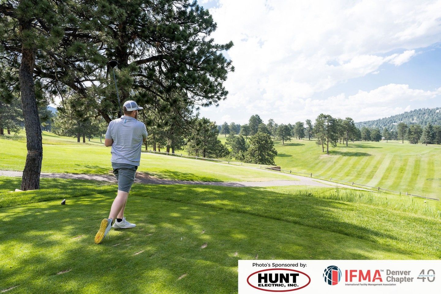 Man teeing off on a golf course under a blue sky, trees in the background.