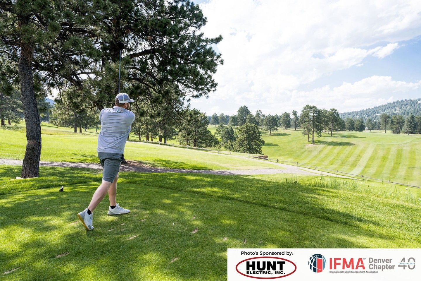 Man swinging golf club on green, tree-lined course under a sunny sky.