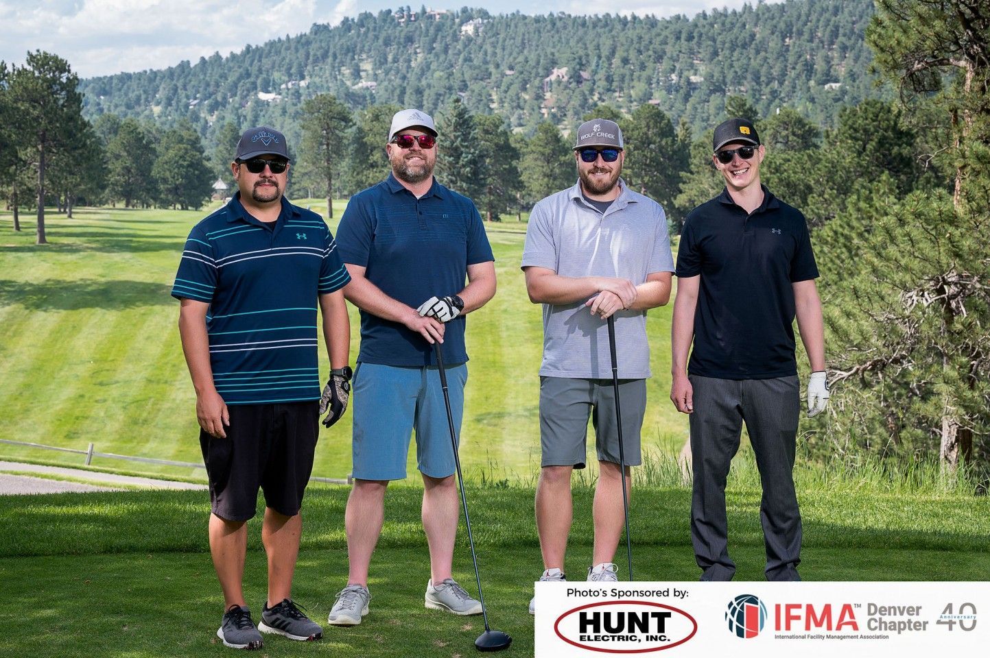 Four men posing on a golf course, mountains in the background, all wearing hats.