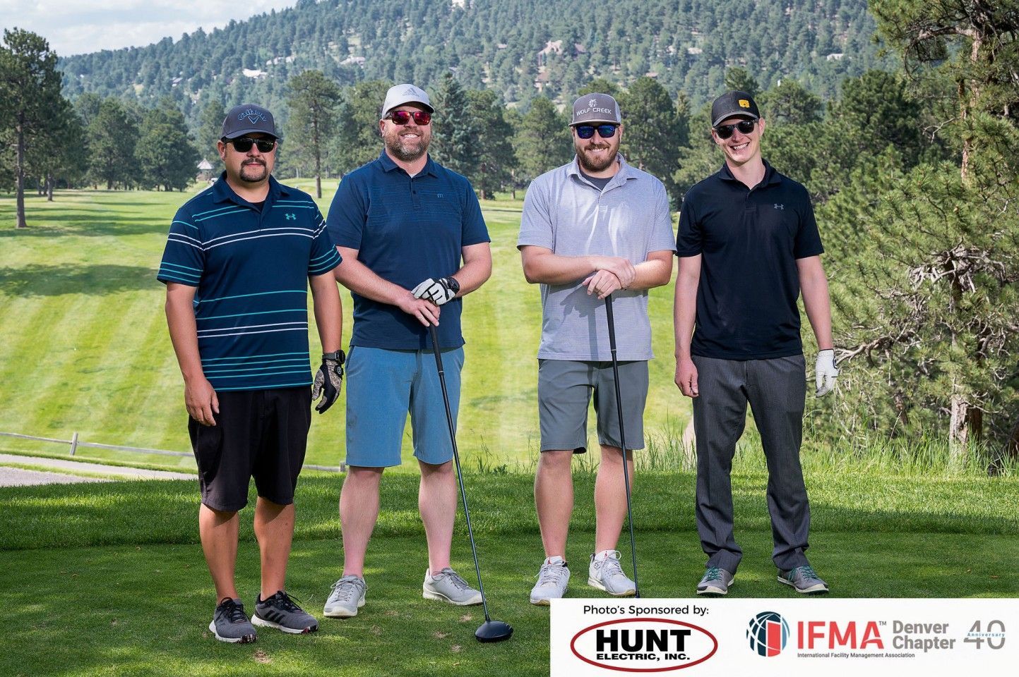 Four men on a golf course pose for a photo, wearing hats and sunglasses. Mountain backdrop.