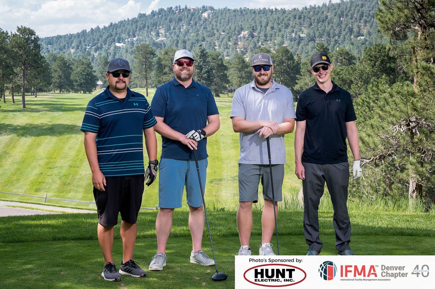 Four men on a golf course. They are wearing sunglasses and holding golf clubs. Background is trees and a hill.