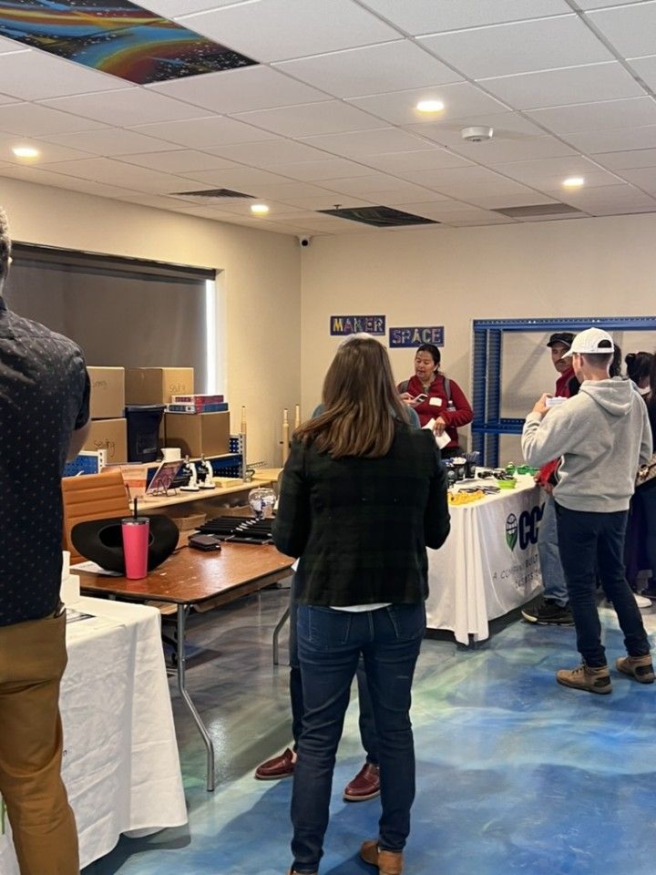 People at an event. Tables with items. A person in a red shirt gestures. Blue and white floor.