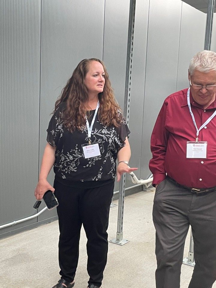 Woman in black top gestures, talking to a man in red shirt. They wear lanyards, in an industrial setting.